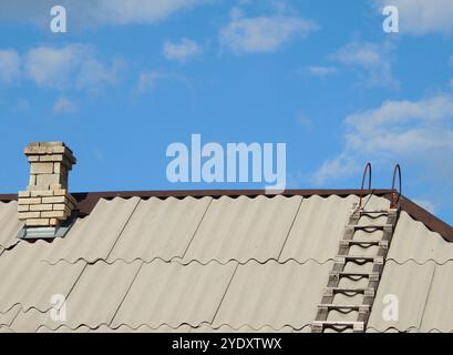 tubo in mattoni di camino e scala in legno sul tetto in ardesia ondulato grigio di una casa rurale privata sotto il cielo azzurro, in cima a un edificio rustico con scale Foto Stock