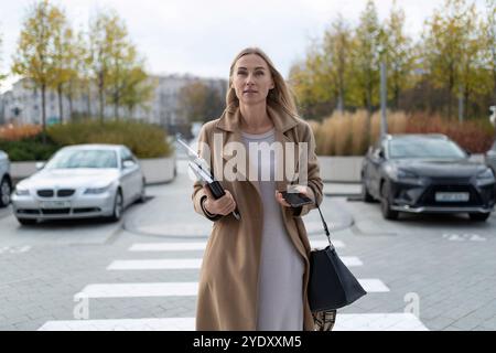 Una donna d'affari cammina vicino a un business center, portando con sé un computer portatile e un telefono, con un aspetto sicuro e pronto per la giornata Foto Stock