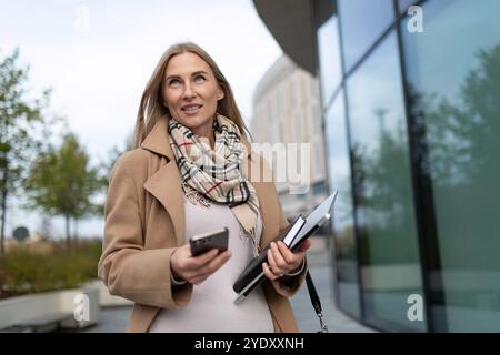 Una donna d'affari cammina vicino a un business center mentre tiene in mano uno smartphone e documenti Foto Stock