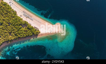 Le onde si infrangono dolcemente contro la riva di Zlatni Rat, dove la vegetazione lussureggiante incontra le acque turchesi. La luce del sole si riflette sulla spiaggia incontaminata, creando un'atmosfera serena per il relax e l'esplorazione. Foto Stock