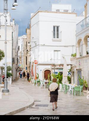 Ostuni Italia 18 settembre 2024 Un residente locale vaga per i pittoreschi vicoli della Puglia, circondato da edifici imbiancati e vivaci caffetterie all'aperto Foto Stock