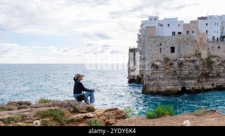 Il visitatore siede tranquillamente su una sporgenza rocciosa, guardando la costa mozzafiato della Puglia e il tranquillo Mare Adriatico, circondato dall'affascinante architettura della città storica. Polignano a Mare Italia Foto Stock