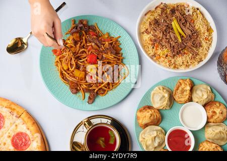 Il tavolo è pieno di piatti e cibo. Set di pilaf uzbeki, lagman, manti. La mano di una donna con una forchetta. Background alimentare. Foto Stock