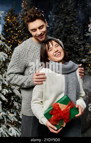 Una coppia felice con maglioni grigi caldi si erge tra abeti innevati, condividendo allegria e amore festivi. Foto Stock