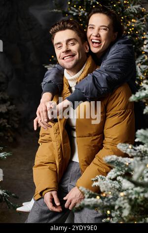 Una coppia felice abbraccia in giacche accoglienti, circondate da pini innevati, catturando la gioia delle vacanze. Foto Stock