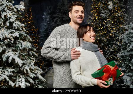 Questa deliziosa coppia abbraccia lo spirito natalizio, circondato da abeti innevati e decorazioni festose. Foto Stock