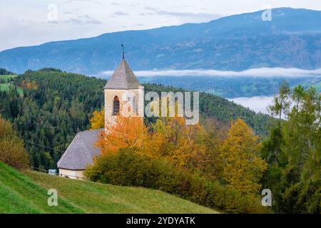 Incastonata nelle pittoresche Dolomiti, una chiesa storica si erge in mezzo a un vivace fogliame autunnale, con la nebbia che si innalza dolcemente attraverso la valle all'alba, creando un'atmosfera serena e incantevole. Foto Stock