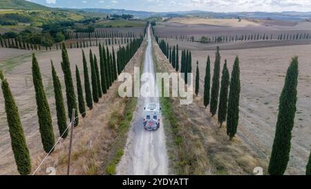 Viaggia lungo la tranquilla strada sterrata incorniciata da maestosi cipressi, catturando l'essenza di un tranquillo paesaggio toscano bagnato dalla luce soffusa del sole, evocando un senso di vagabondaggio e avventura. Foto Stock