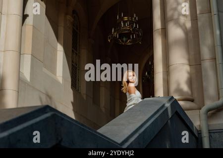 Donna, scale, edificio - donna bionda si erge su gradini di pietra fuori dall'ingresso di un edificio con la luce del sole che splende dentro. Foto Stock