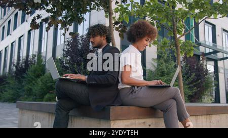 Due persone colleghi multirazziali Diversity partner commerciali che lavorano all'aperto su una panchina con due laptop donna africana uomo indiano arabo lavoro Foto Stock