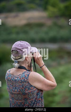 una donna anziana o di mezza età che fa birdwatching guardando attraverso un binocolo con un berretto da baseball. Foto Stock