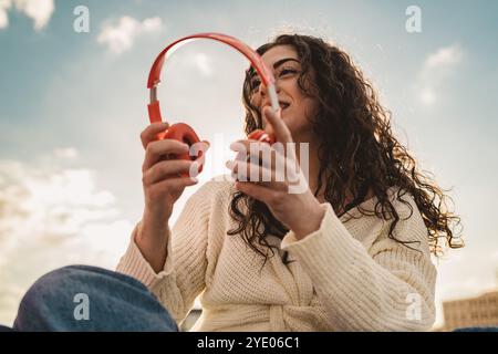 Bella donna variegata con capelli ricci naturali e bretelle che ascoltano musica all'aperto. Ritratto di stile di vita moderno che mostra l'autentica cultura giovanile e se stessi Foto Stock