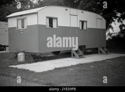 Anni '1950, storica, una carovana festiva dell'epoca, parcheggiata su un pavimento di cemento in un campo, scalini di legno fino a due porte d'ingresso laterali, Inghilterra, Regno Unito. L'aumento del numero di persone che possedevano automobili negli anni '1950 e '60 ha fatto sì che le famiglie ora potessero viaggiare e utilizzare tali roulotte per le vacanze, noleggiate per una settimana o possedute, con una frequenza molto più elevata di prima. La pubblicità li ha promossi come un essere "omobile", letteralmente, una casa su ruote. Foto Stock