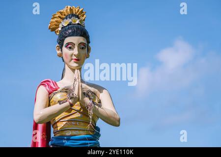 Statua colorata di donna balinese che accoglie i visitatori con le mani unite in preghiera, adagiata contro un cielo blu brillante al tempio Tanah Lot di Bali Foto Stock