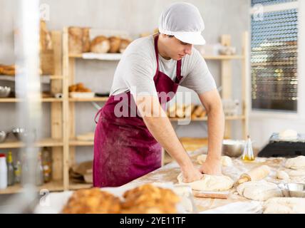 Giovane panettiere positivo in grembiule impastare e formare l'impasto sul tavolo, processo di preparazione del pane in cucina Foto Stock
