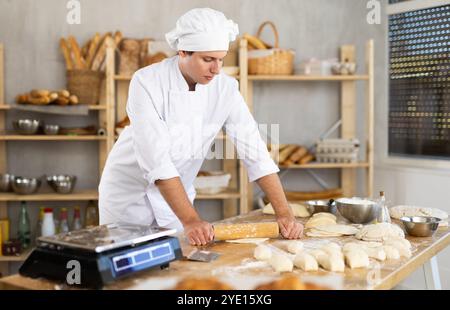 Giovane chef con uniforme bianca che tiene in mano il mattarello e stende l'impasto nella cucina dei panifici Foto Stock