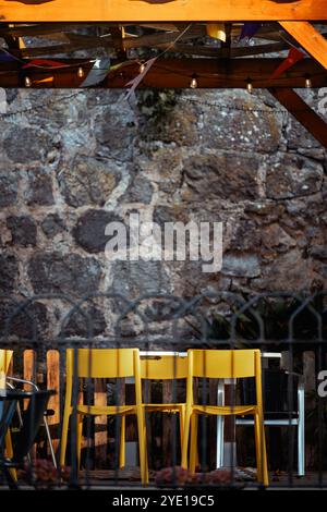 Sedie gialle luminose disposte sotto un pergolato di legno adornato da luci e colorati nastri sospesi. Il muro di pietra sullo sfondo aggiunge rustico te Foto Stock