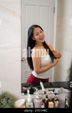 Una bella e felice donna asiatica con lunghi capelli neri naturali si pettina i capelli con una spazzola di legno dopo la doccia, sorridendo al suo riflesso Foto Stock