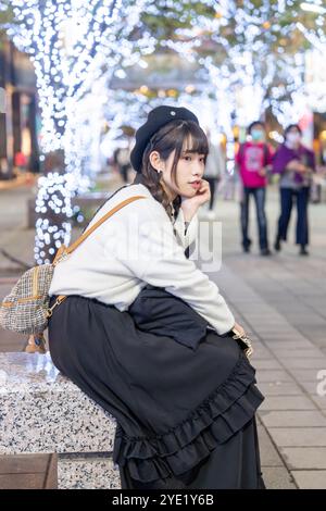 Una giovane e carina donna taiwanese di 20 anni che indossa abiti in bianco e nero, seduta e riposante di notte in mezzo a splendide illuminazioni in uno shopping Foto Stock