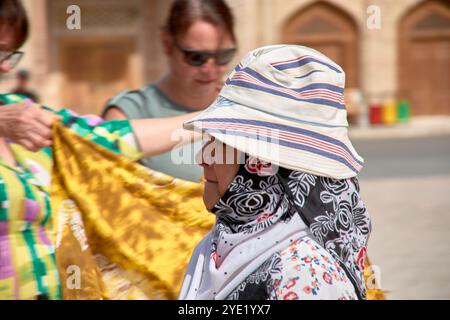 Bukhara, Uzbekistan; 19 settembre 2024: Primo piano di un'anziana donna uzbeka che indossa un cappello tradizionale, sullo sfondo di un vivace marchio di seta Foto Stock