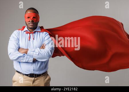 serio uomo afroamericano in costume da supereroe che piega le braccia isolate su sfondo bianco. ragazzo dalla pelle scura con maschera rossa e mantello, super power co Foto Stock