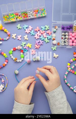 La bambina ha fatto braccialetti su sfondo viola. Gioielli con perline fatti a mano per bambini. Collane e bracciali realizzati con perle e perle multicolore. Kit fai da te Foto Stock