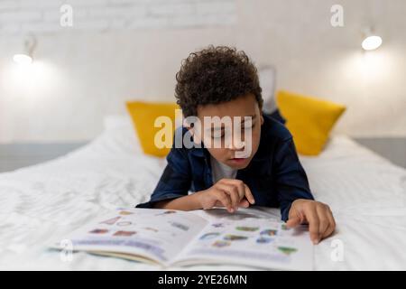 Ragazzo afroamericano dai capelli ricci sdraiato sul letto con un libro e sembra eccitato Foto Stock