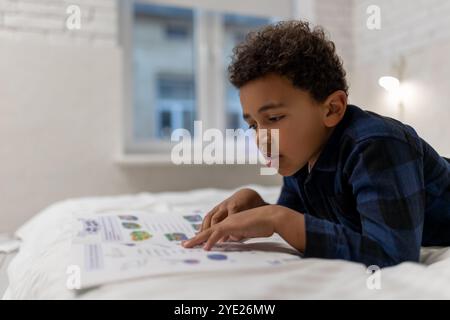 Ragazzo afroamericano dai capelli ricci sdraiato sul letto con un libro e sembra eccitato Foto Stock