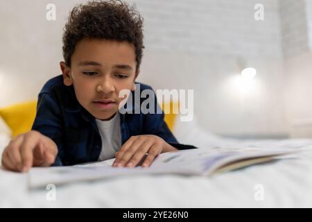 Ragazzo afroamericano dai capelli ricci sdraiato sul letto con un libro e sembra eccitato Foto Stock