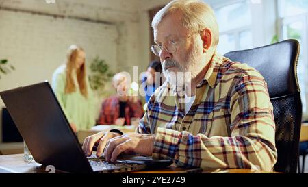 Uomo anziano, professionista, seduto in uno spazio di lavoro collaborativo, alla scrivania con notebook che ricerca, esamina report o analisi. Foto Stock