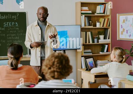 Insegnante esperto che utilizza un tablet, conduce discussioni di gruppo in classe con gli studenti che ascoltano attentamente mentre presentano le loro presentazioni su un televisore a schermo piatto Foto Stock