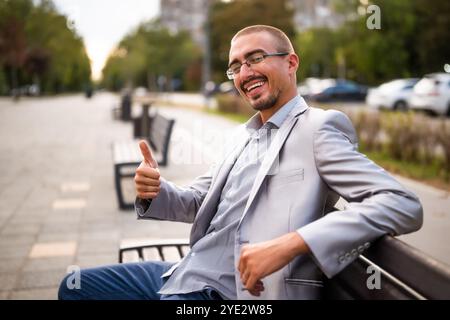 Ritratto di un allegro uomo d'affari seduto sulla panchina della città. Uomo felice che guarda la macchina fotografica e sorride. Foto Stock