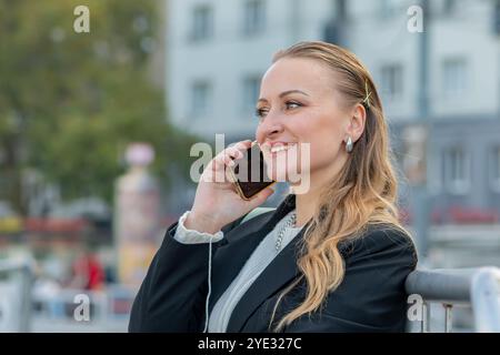 Una donna di mezza età di 40-45 anni che parla al telefono mentre si siede su una panchina in una città vivace, trascorrendo una piacevole giornata con un sorriso sul viso. Foto Stock