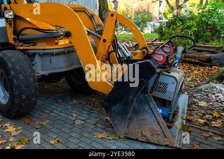 Un caricatore giallo con benna nera è parcheggiato su un vialetto di mattoni. La benna viene sollevata, rivelando una piastra vibrante grigia dietro di essa. Foto Stock