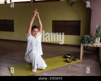 Donna in abiti bianchi che fa esercizio Sat kriya, innalzando l'energia sulla colonna vertebrale, toccando il pavimento con un ginocchio e le braccia estese verso l'alto, esercitando K Foto Stock