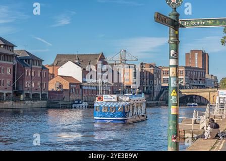 Un tour in barca scivola lungo un fiume con magazzini convertiti sulla riva lontana. Un ponte è in lontananza e un cartello è in primo piano. Foto Stock