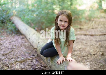 Una bambina con una maglietta verde si siede nella foresta su un tronco di albero caduto. Il concetto di unità con la natura e l'infanzia Foto Stock