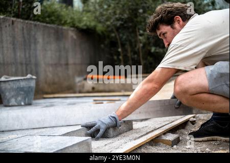 Un uomo che indossa guanti e si concentra sul lavoro, allinea una livella metallica su mattonelle di blocchi di pietra in un'area di costruzione all'aperto. Foto Stock