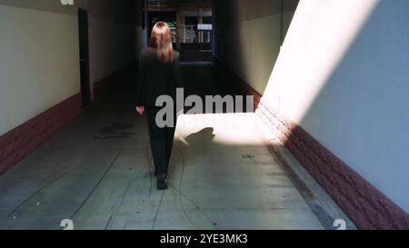 Una giovane donna con una tuta classica nera cammina con sicurezza attraverso un arco della casa, con il gioco di ombre e la facciata della casa che crea un effetto sorprendente Foto Stock