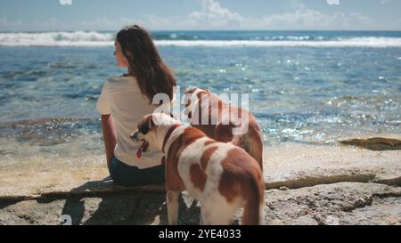La donna si siede con i suoi cani vicino al mare, godendosi la vista in una giornata di sole. Sembrano felici e rilassati, creando una tranquilla scena di amicizia Foto Stock