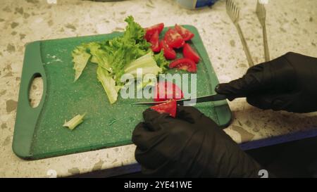 Le verdure tritate giacciono su una tavola verde. La ragazza chef con i guanti neri taglia un pomodoro. Foto Stock