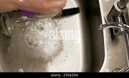 Un primo piano di mani che lava un recipiente in acciaio inox sotto l'acqua corrente nel lavandino. L'attenzione è rivolta al processo di pulizia e alla cucina quotidiana Foto Stock