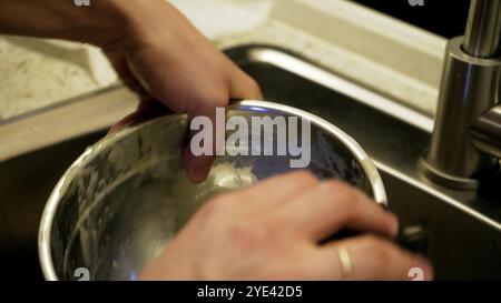 Un primo piano di mani che lava un recipiente in acciaio inox sotto l'acqua corrente nel lavandino. L'attenzione è rivolta al processo di pulizia e alla cucina quotidiana Foto Stock