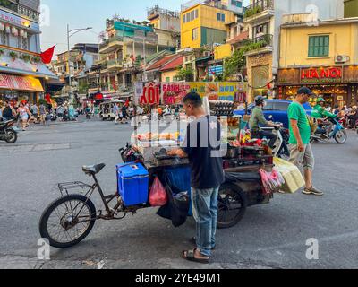 Hanoi, Vietnam: Un vietnamita che cucina il cibo sul suo carrello a ruote in mezzo alle strade affollate della città, alle auto e alle moto che passano a tutta velocità Foto Stock