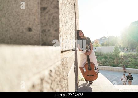 immagine a figura intera di una giovane donna latina appoggiata su un muro con una chitarra acustica in mano. vista sullo sfondo della città con il sole che splende Foto Stock