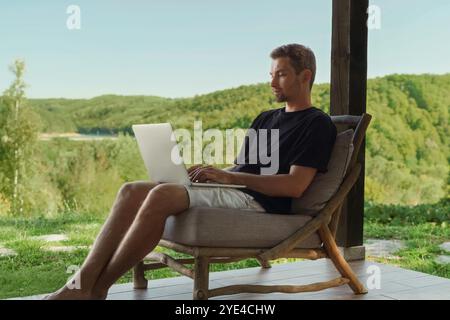 Il libero professionista lavora online sulla terrazza di una casa di campagna, godendo della natura. Lavoro a distanza e equilibrio della vita privata Foto Stock