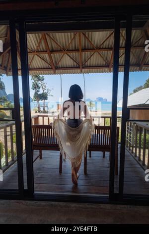 Una giovane donna si erge graziosamente su un ponte di legno, drappeggiato in uno scialle leggero. Guarda la spiaggia tranquilla e le isole lontane durante l'ora d'oro, e. Foto Stock