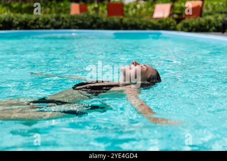 Una giovane donna gode di un momento di tranquillità, galleggiando in una piscina rinfrescante sotto il sole luminoso, circondata da una vegetazione vivace e sedie a sdraio. Foto Stock