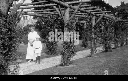 West Sussex, Inghilterra. c.1913 - Una donna e un giovane ragazzo, che si ritiene siano parenti della famiglia Henty, si trovano sotto un pergolato rustico all'interno dei terreni di Ferring Grange, situato a Ferring, un villaggio costiero nel West Sussex. Questa tenuta era la casa di Edwin Henty, J. P, D.L., F.S.A. (1844 – 1916), che era stato alto sceriffo del Sussex. Nel 1924, la casa fu convertita in un hotel alla moda, visitato da molte celebrità tra cui Edoardo, il Principe di Galles (in seguito noto come Duca di Windsor). La casa fu distrutta da un incendio nell'ottobre 1946. Foto Stock