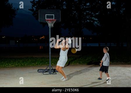 Un padre gioca a basket con il suo giovane figlio di notte in un parco illuminato da una luce brillante Foto Stock